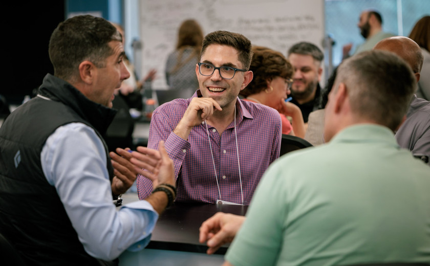 A photo of two math educators—one facing the camera, one with his back to it—listen as a third—sitting sideways—animatedly explains something to them.