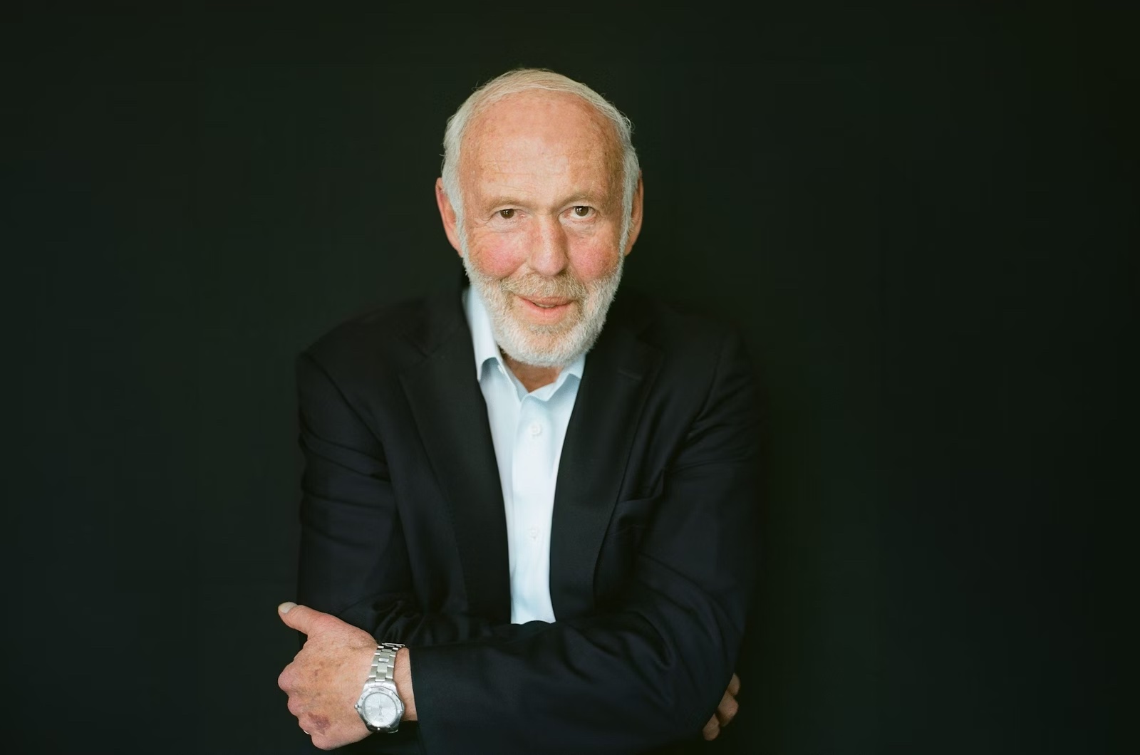 An elderly man with white hair and a short beard wearing a light blue shirt and black jacket looking at the camera with his arms crossed.