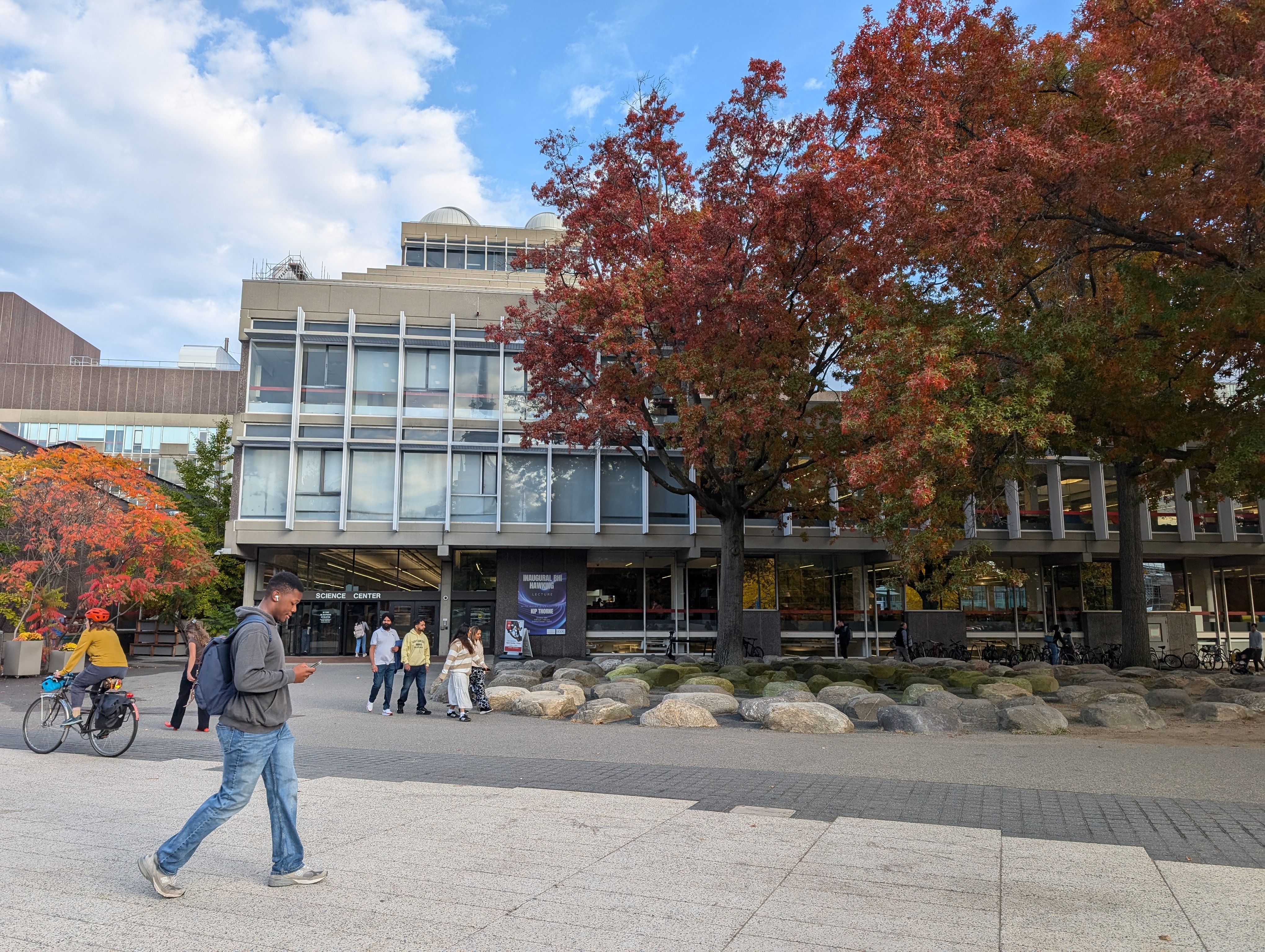 A photo of the Harvard Science Center building with fall foliage and students walking across the plaza.