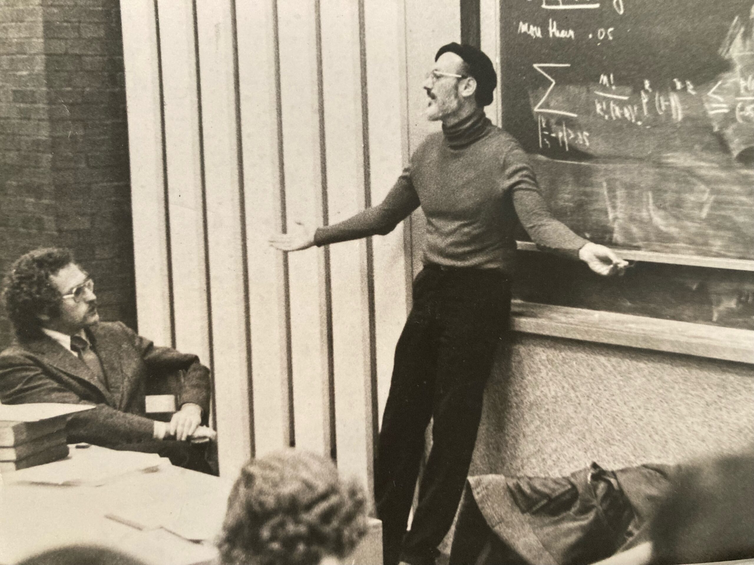 A black and white photo of Harvard mathematician Shlomo Zvi Sternberg in the 1970s as he gives a lecture leaning against a blackboard with math formulae.