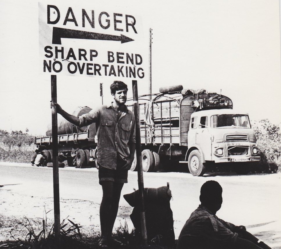 A 1972 black-and-white image of Harvard math professor emeritus Dick Gross hitching across Kenya, standing under a traffic sign.